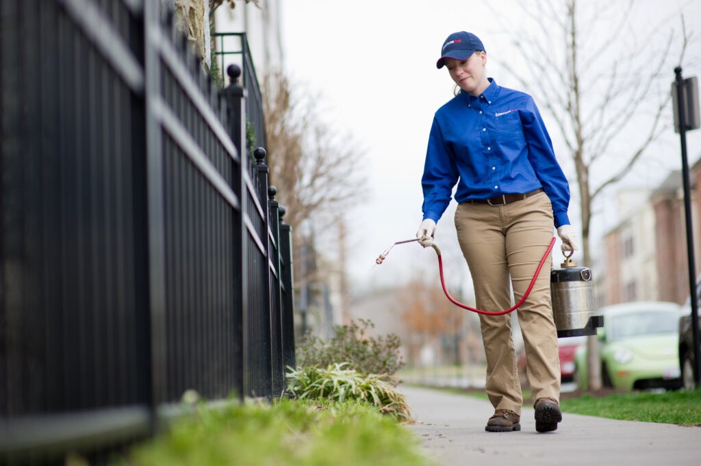An American Pest technician carrying exterior treatment equipment along a sidewalk